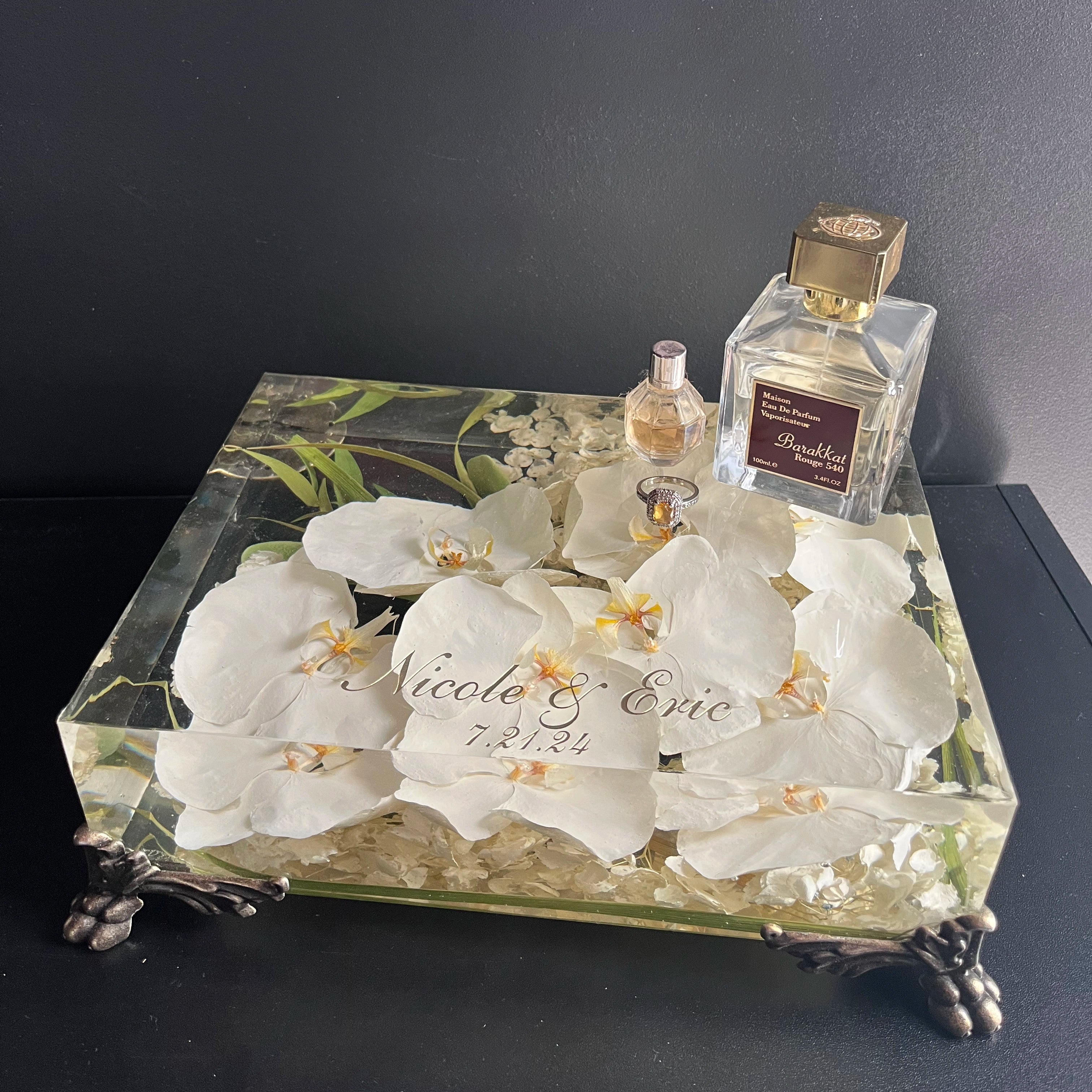 Decorative box with white flowers and engraved text, accompanied by perfume bottles on a dark surface.