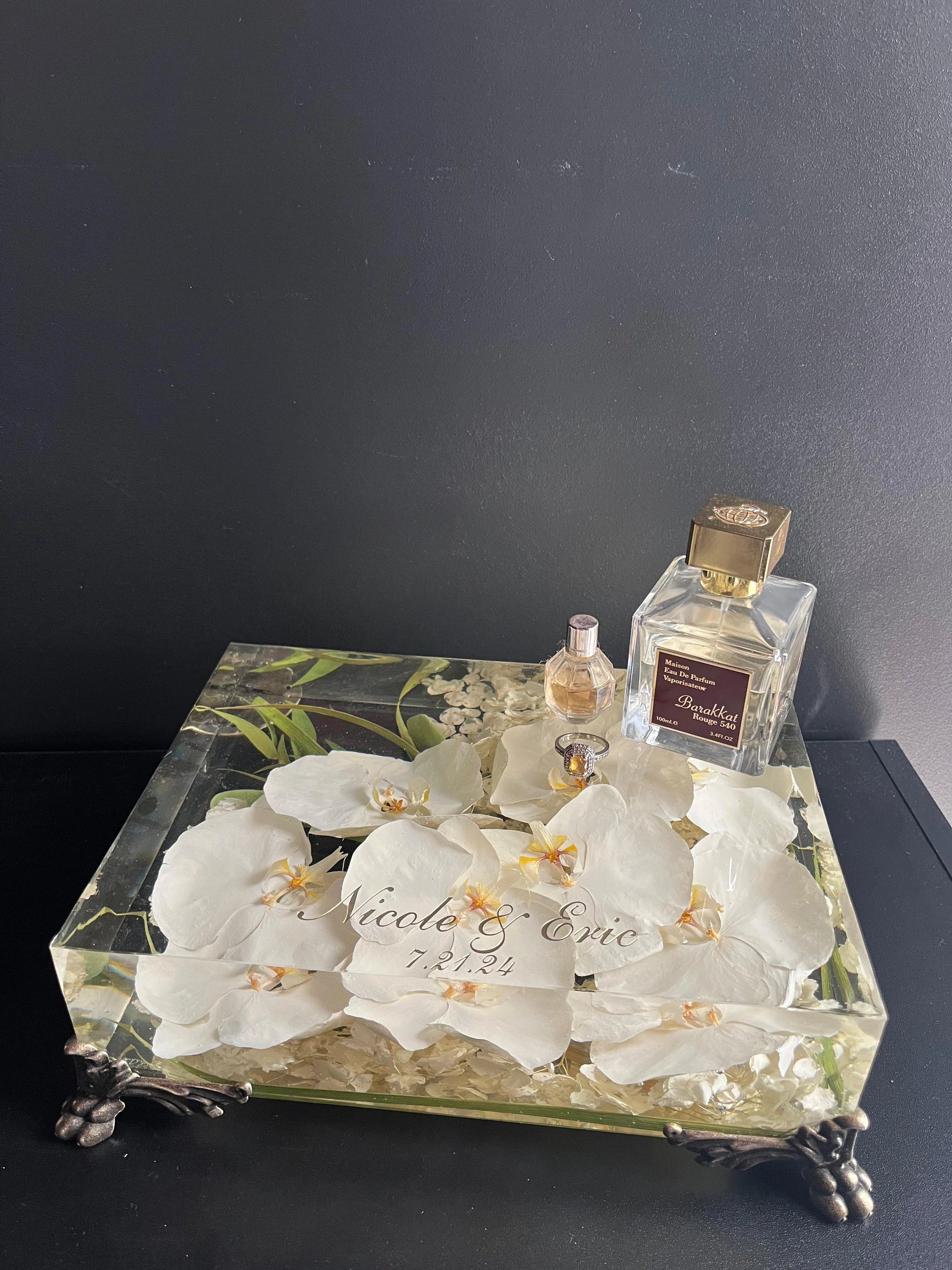 Decorative box with white flowers and engraved text, accompanied by a perfume bottle on a dark surface.